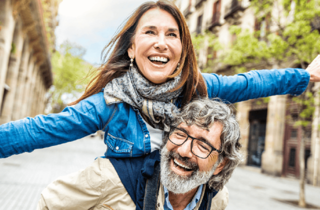 Joyful older couple enjoying a piggyback ride on a lively city street lined with trees and historic buildings.