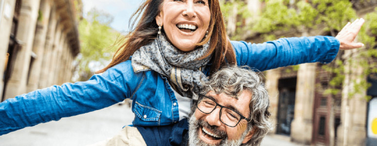 Joyful older couple enjoying a piggyback ride on a lively city street lined with trees and historic buildings.