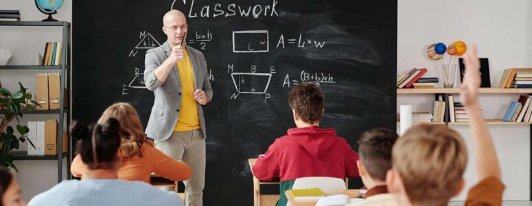 Middle school teacher standing at the front of a classroom of students in front of a chalkboard displaying classwork