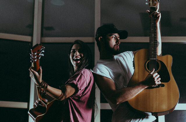 Two people on stage, both playing the guitar and one signing, in front of a window
