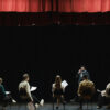 Actor standing on a stage in front of four students who are sitting and holding scripts