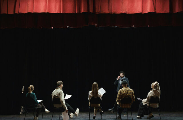 Actor standing on a stage in front of four students who are sitting and holding scripts