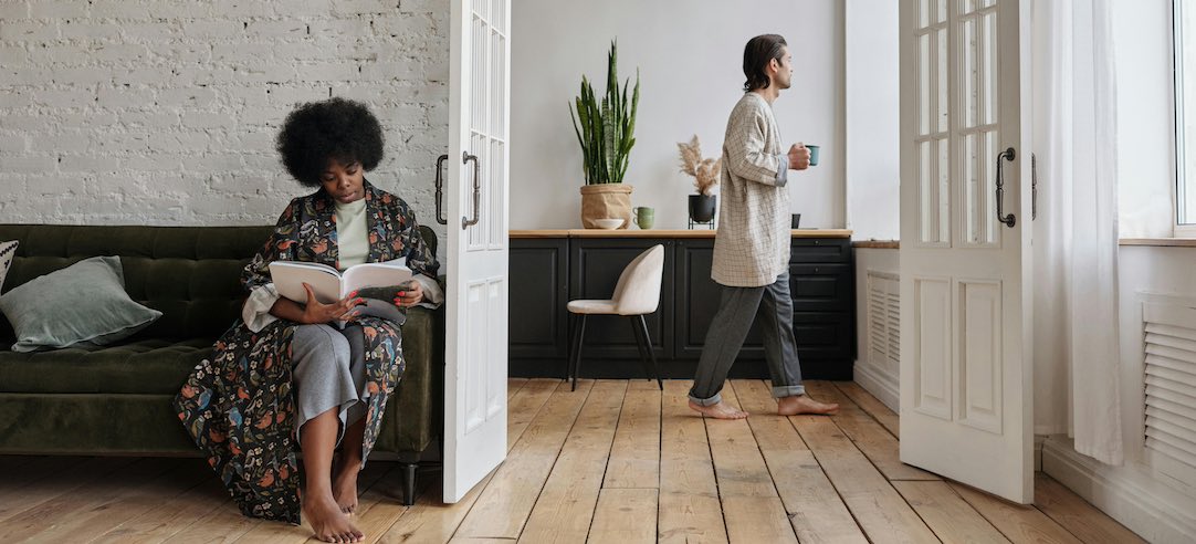 Woman sitting on couch reading while her partner looks out the window.