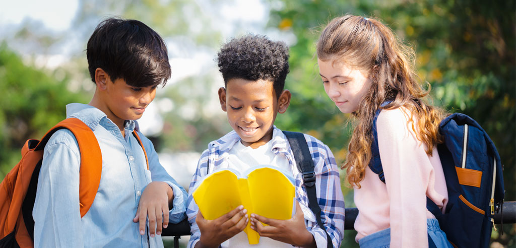 Elementary-aged children standing outside and looking at a yellow book together