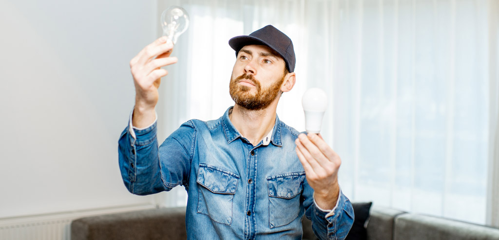 Man sitting on his couch in his apartment trying to save money by swapping out an expensive lightbulb for a cheaper one