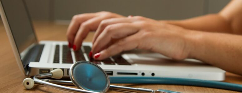 Close view of a nurse writer's hands on their laptop with a stethoscope sitting next to it.
