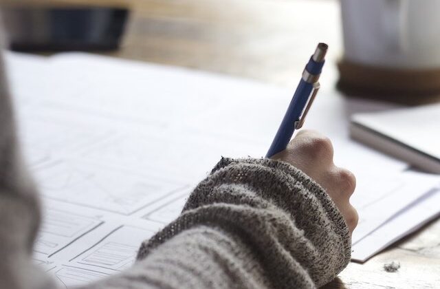 Close view of a writer at their desk holding a pen and working on an article.