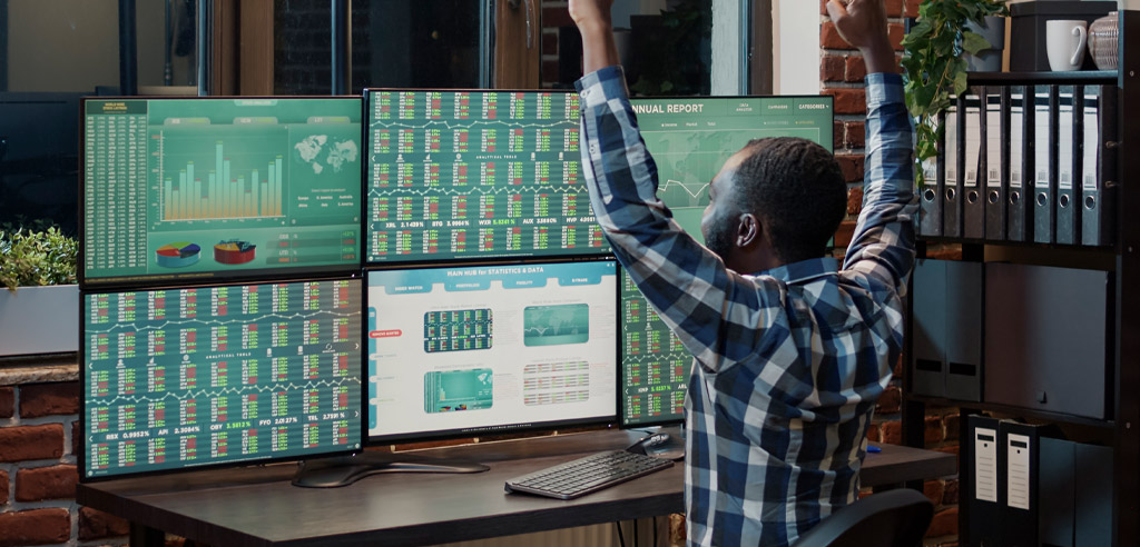 Man sitting at a desk looking at a display of his investments with his arms in the air