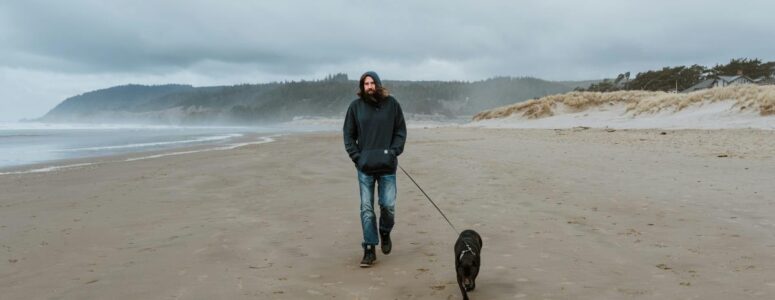 Professional dog walker wearing a hoodie and walking along the beach with a medium-sized black dog on a leash