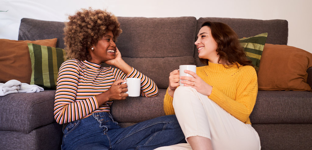 Two women sitting against a couch sipping drinks and talking about their monthly budget