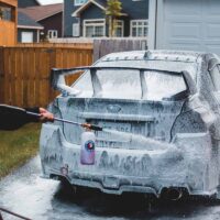 Car wash business owner hosing down a car in a driveway.