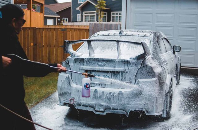 Car wash business owner hosing down a car in a driveway.