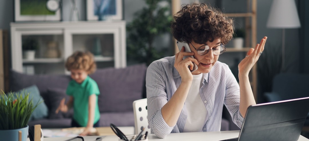 Mother answering a work call from home while her child plays in the background.