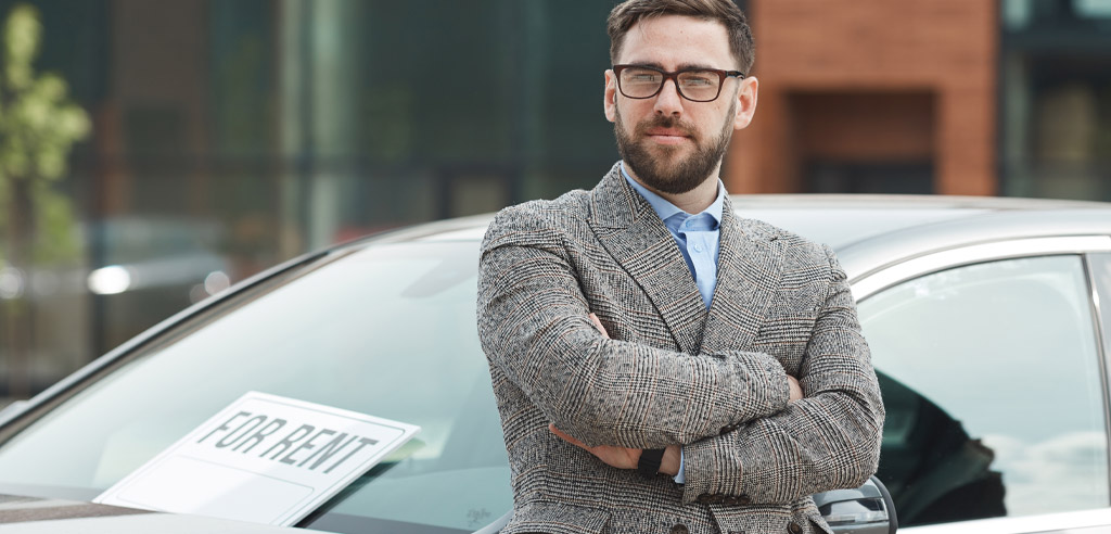 Man with crossed arms standing in front of a car with a For Rent sign on the windshield