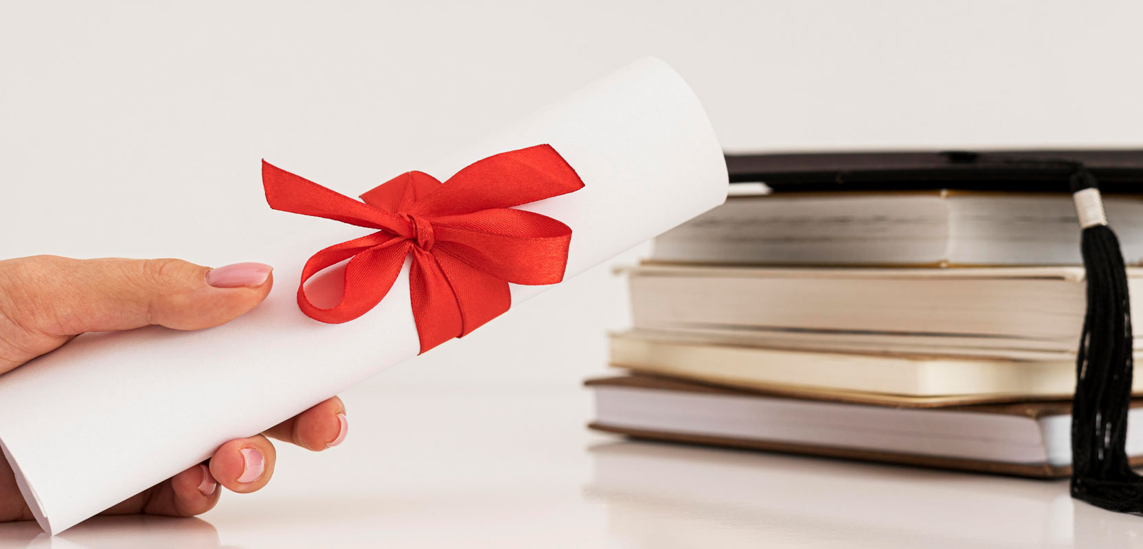 College diploma wrapped up with a ribbon next to a pile of books and a traditional graduate's cap