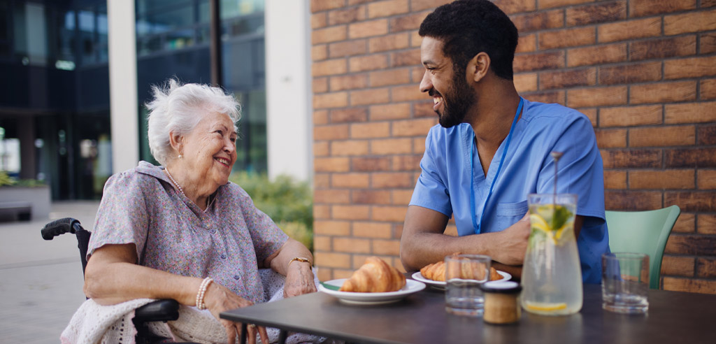 Male caregiver sitting with an older woman at a table with food as part of his paid caregiving side hustle