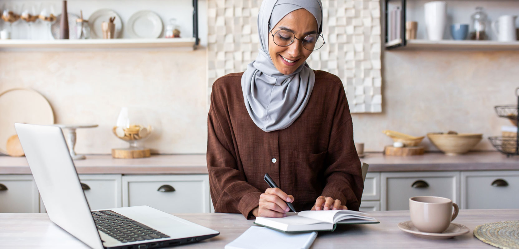 Freelance writer sitting in her kitchen taking notes in a notebook next to her laptop