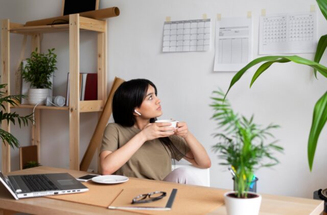 Woman at desk drinking coffee and looking at a wall calendar.